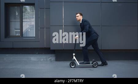 lunghezza completa di uomo d'affari in occhiali e tuta da guida e-scooter vicino all'edificio, immagine stock Foto Stock