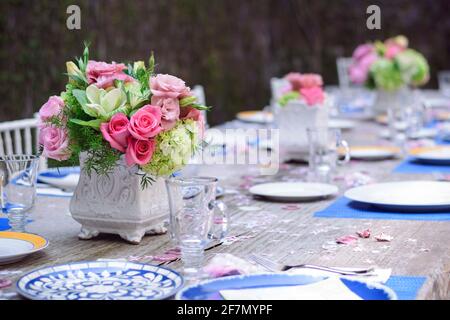 Tavolo in giardino con piatti in porcellana blu e gialla, con vetri di cristallo vuoti e una disposizione floreale al centro con rose rosa e bianco Foto Stock