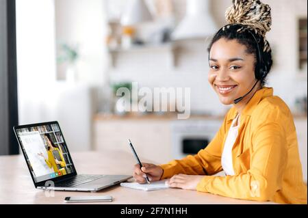 Apprendimento a distanza tramite videochiamata. Happy African American studentessa indossa le cuffie, si siede alla scrivania a casa, studia da remoto, guarda la fotocamera, sorriso amichevole, insegnante femminile sullo schermo del computer portatile Foto Stock