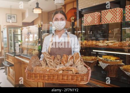 Panettiere femminile che indossa una maschera medica per il viso, con cestino pieno di pane appena sfornato Foto Stock