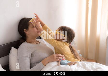 bambina che indossa un medico che gioca in uniforme, infermiera con mamma giovane, pannaia in camera da letto, controllo della gola della madre, misura la temperatura, si prenda cura, famiglia Foto Stock