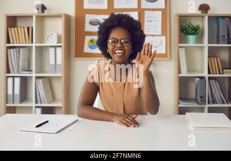 Donna afro-americana che agita la mano guardando la macchina fotografica seduta a. scrivania da ufficio Foto Stock