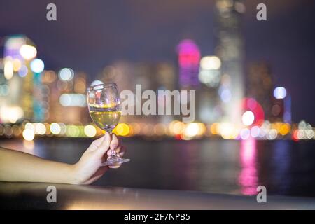 lady Hand tiene due bicchieri di vino con luci notturne della città sfocate e scintillanti nel centro, Hong Kong Victoria Harbour come sfondo, concetto romantico per Foto Stock