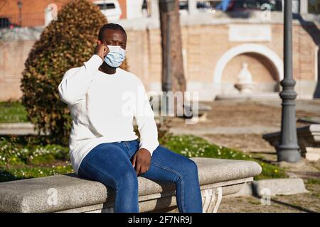 Giovane afroamericano seduto su una panchina del parco che parla al telefono. Uomo latino con maschera anti-contagio. Concetto di nuova normalità. Foto di alta qualità Foto Stock