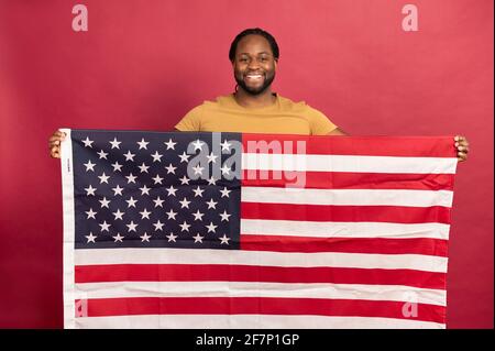 Un uomo nero e muscoloso sorrise che tiene davanti a sé la bandiera americana, indossando una t-shirt in senape, in piedi al coperto su sfondo rosso, mostra la sua indipendenza e i diritti di una persona uguale Foto Stock