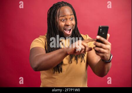 Eccitato sorridente uomo scuro skinned con lunghi Dreadlock che tengono il telefono, leggendo buone notizie nel messaggio, guardando lo schermo, celebrando la vittoria della lotteria in linea, scorrendo le informazioni, su sfondo rosso Foto Stock