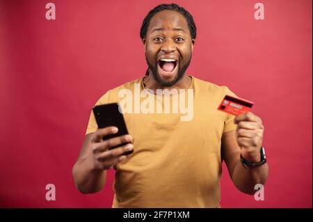 Felice uomo africano sopravorato tenendo in mano la carta di credito e il cellulare, in piedi su uno sfondo rosso con bocca aperta, comodamente facile e veloce da pagare utilizzando l'applicazione sul telefono cellulare Foto Stock