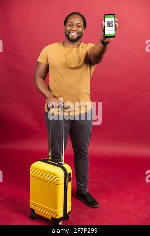 Uomo africano felice in piedi con la piccola borsa e cellulare alla moda in mano, utilizzando il codice QR per il controllo di passaggio veloce e sicuro e dogana, avendo un biglietto online, viaggiare con concetto di comfort Foto Stock