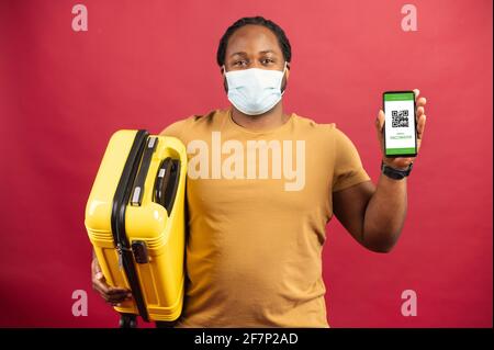 Uomo africano felice in maschera medica in piedi sullo sfondo rosso che tiene la borsa gialla per il viaggio e cellulare con codice QR sullo schermo, concetto di esposizione biglietto senza contatto, passaggio sicuro della dogana Foto Stock