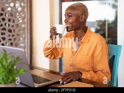 Donna anziana africana che beve caffè mentre usa il computer portatile al bar Ristorante - concetto di lavoro intelligente Foto Stock