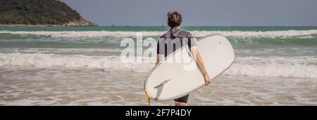 STRISCIONE, LUNGO FORMATO giovane surfer in piedi contro l'oceano guardando le onde, surfer professionale in muta nera tenendo con una mano grande bello Foto Stock