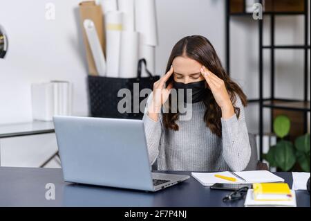 Stanco giovane donna d'affari che indossa maschera medica protettiva sentire sintomi di malattia virale, ha mal di testa, seduto sul posto di lavoro in ufficio, tiene e massaggiare templi con sofferenza espressione del volto Foto Stock