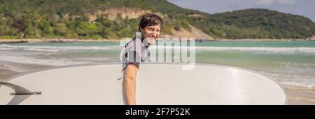 STRISCIONE, LUNGO FORMATO giovane surfer in piedi contro l'oceano guardando le onde, surfer professionale in muta nera tenendo con una mano grande bello Foto Stock