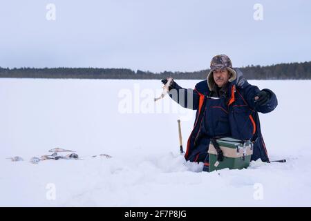 pesca sul ghiaccio. pesca del pescatore su un lago d'inverno contro uno sfondo di foresta e cielo blu. Foto Stock