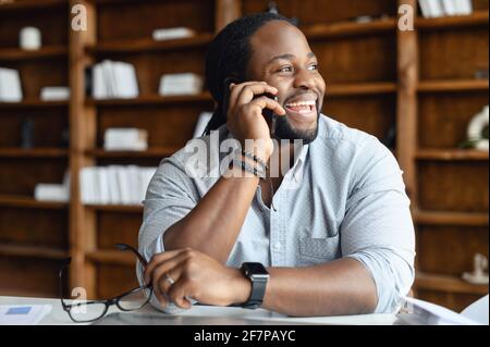 Allegro afroamericano maschio dipendente che indossa una camicia casual intelligente che tiene conversazione telefonica, un uomo seduto alla scrivania in un ufficio moderno, un imprenditore biraciale che parla di smartphone e ride Foto Stock