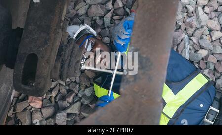 Un tecnico africano di macchina che si sdraiava e indossa un casco, un boschetto e un giubbotto di sicurezza sta usando una chiave per riparare il treno Foto Stock