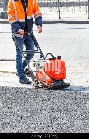 Un operatore su strada in una giacca riflettente arancione brillante utilizza una piastra vibrante per compattare l'asfalto in un luogo di riparazione stradale. Immagine verticale, spazio di copia. Foto Stock