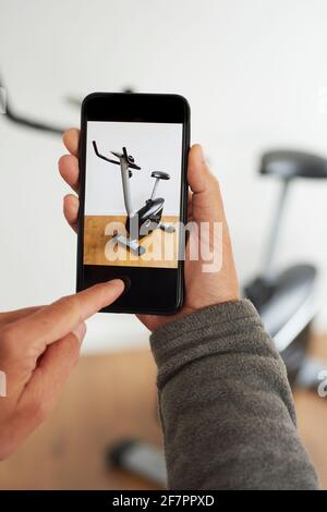 un giovane caucasico scatta una foto, con il suo smartphone, di una bicicletta stazionaria per venderla su un'app per il mercato online per vendere e acquistare la seconda mano Foto Stock