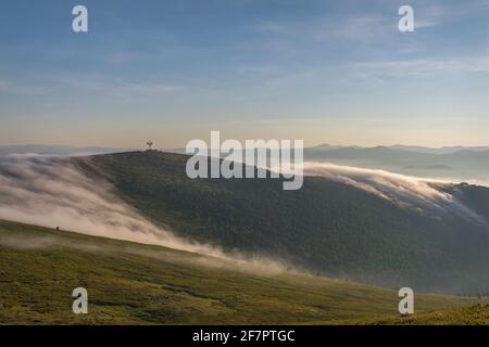 Incredibile alba con bella nebbia soffusca, torre di telecomunicazioni in cima alla montagna e foresta di cedri sullo sfondo di cielo blu e cl Foto Stock