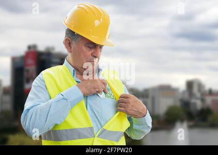 Vecchio costruttore maschio che indossa il cappello e le attrezzature da lavoro nascondono i contanti stipendio denaro nella tasca del petto della camicia come lavoro di occupazione concetto Foto Stock