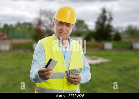 Vecchio costruttore maschile indossando gilet fluorescente e supporto giallo hardhat denaro in contanti come stipendio e pagamento con carta di credito o debito concetto di retribuzione Foto Stock