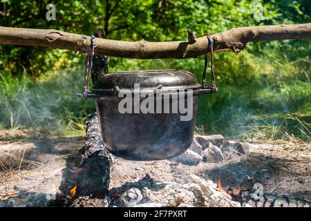 Una pentola per cucinare all'aperto è appesa su un bastone. I tronchi sbodano sotto e il fumo è visibile. Lo sfondo è sfocato. Cucina campo Foto Stock