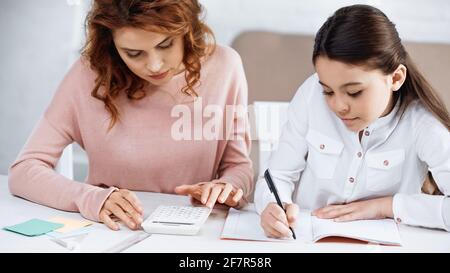 Ragazza che scrive sul notebook vicino a madre e calcolatrice a casa Foto Stock