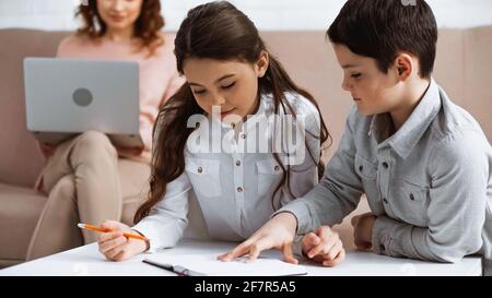 Fratelli con matita e notebook vicino alla madre utilizzando il computer portatile su sfondo sfocato a casa Foto Stock