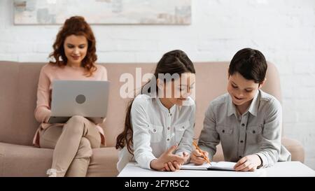 Sorridendo i bambini che scrivono sul notebook vicino alla madre utilizzando il computer portatile sfondo sfocato nel soggiorno Foto Stock