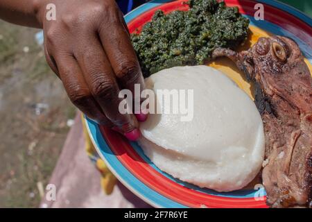 Servendo a mano su un piatto contenente cibo di base di panino africano, stufato di foglie di zucca e maiale alla griglia Foto Stock