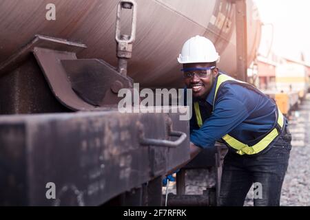 Il tecnico africano di macchina che indossa un casco, boschetti e giubbotto di sicurezza sta usando una chiave per riparare il gas e l'olio di trasporto del treno Foto Stock
