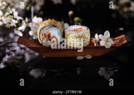 Due rotoli di tempura con riso, formaggio e pesce in una barca decorativa in legno su sfondo nero. Cucina giapponese Foto Stock