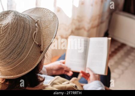 Donna anziana con un cappello sopra, leggendo un libro vicino alla finestra. Luce pomeridiana Foto Stock