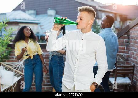 Giovane ed elegante che beve birra dalla bottiglia durante il party sul tetto. Tre amici diversi che chiacchierano e sorridono sullo sfondo. Weekend spensierati di giovani. Foto Stock