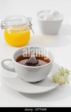 Una tazza di tè caldo viene prodotta da una bustina di tè. Fiori su un piatto, miele e zucchero sullo sfondo. Profondità di campo poco profonda Foto Stock