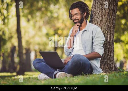 Ritratto di bello allegro viaggiatore focalizzato ragazzo blogger seduta su erba verde utilizzando un computer portatile che lavora una giornata di sole all'aperto Foto Stock