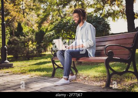 Profilo vista laterale ritratto di allegro viaggiatore focalizzato ragazzo utilizzando computer portatile seduto su panca soleggiata giorno all'aperto Foto Stock