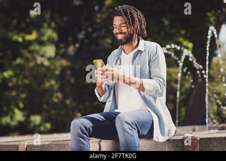 Ritratto di simpatico ragazzo allegro seduto vicino all'acqua su fresco aria utilizzando il dispositivo che chattano i media di navigazione urbano fuori esterno Foto Stock