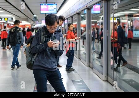 Hong Kong - Novembre, 2019: Giovane uomo che guarda sul telefono cellulare mentre attende il treno alla stazione MTR / stazione della metropolitana di Hong Kong Foto Stock