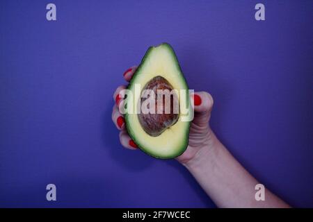 Primo piano della mano di una donna con un dito dipinto di rosso unghie che tengono un avocado a metà taglio contro il dorso viola Foto Stock