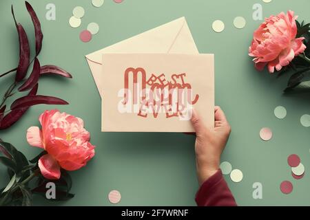 Design biglietto d'auguri per il giorno della mamma. Fiori di peonia rosa su sfondo verde sbiadito, testo migliore mamma mai su carta di carta con busta. Alla moda casual naturale Foto Stock