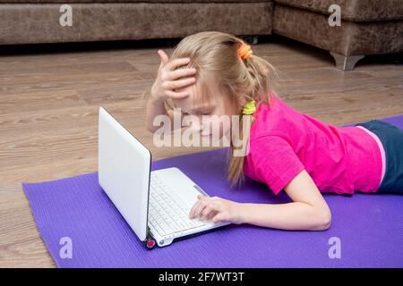 Caucasica ragazza in una T-shirt rosa e jeans si trova sulla stuoia con un computer portatile e preme i tasti. La ragazza si sta preparando per gli sport in linea con un insegnante. Foto Stock