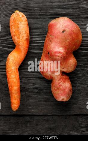 Patate brutto a forma ritorta e carote storte su fondo nero di legno. Concetto di rifiuti vegetali o alimentari, primo piano. Foto Stock