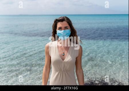 Ritratto di giovane donna in piedi contro l'acqua blu limpida sul mare guardando la macchina fotografica che indossa una maschera chirurgica protettiva per il viso. Foto Stock