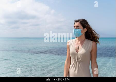 Ritratto di giovane donna in piedi contro l'acqua blu chiaro sopra il mare che guarda via indossando una maschera chirurgica protettiva per il viso Foto Stock