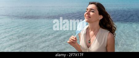 Bandiera orizzontale o ritratto di intestazione di giovane donna sorridente che prende fuori una maschera chirurgica protettiva per respirare aria pura fresca sulla spiaggia. Beautifu Foto Stock