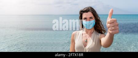 Banner orizzontale o testata Ritratto di giovane donna indossando maschera protettiva che mostra il pollice in alto. Splendido sfondo con acqua blu chiara Foto Stock