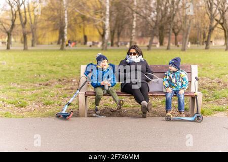 POZNAN, POLONIA - 04 aprile 2021: Donna e due bambini con scooter seduti su una panchina di legno al parco Jan Pawla in un primo giorno di primavera durante Easte Foto Stock