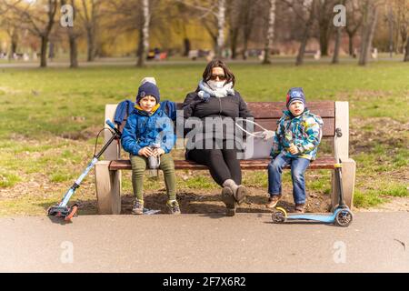 POZNAN, POLONIA - 04 aprile 2021: Donna e due bambini con scooter seduti su una panchina di legno al parco Jan Pawla in un primo giorno di primavera durante Easte Foto Stock