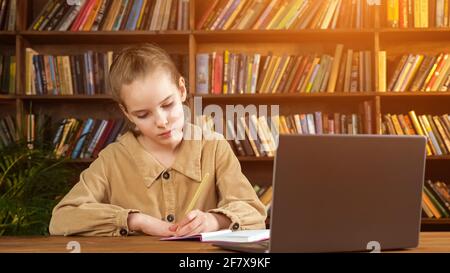 La giovane studentessa in giacca marrone scrive la lezione in carta notebook con matita nella mano sinistra al computer portatile contro grande libreria in legno in biblioteca Foto Stock